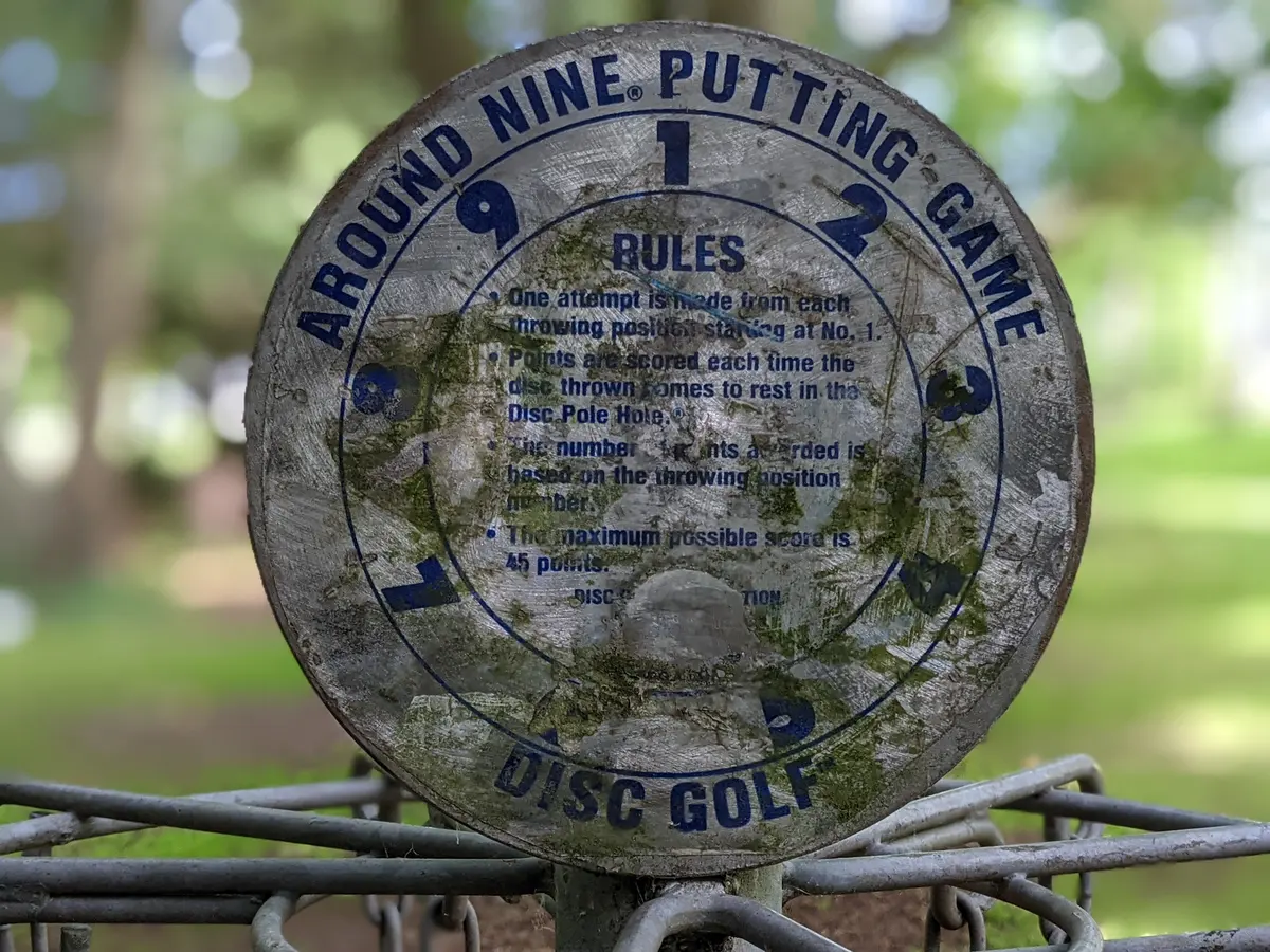 Weathered circular Around Nine Putting Game sign mounted on a disc golf basket